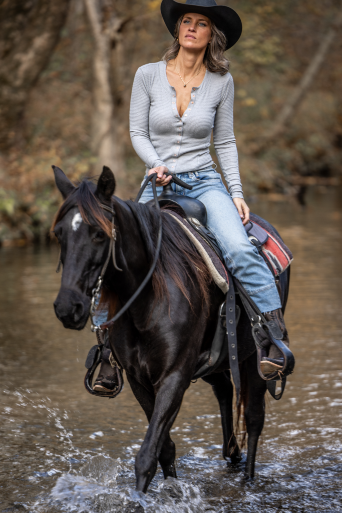 Woman in a cowboy hat riding a black horse through a wooded area in autumn